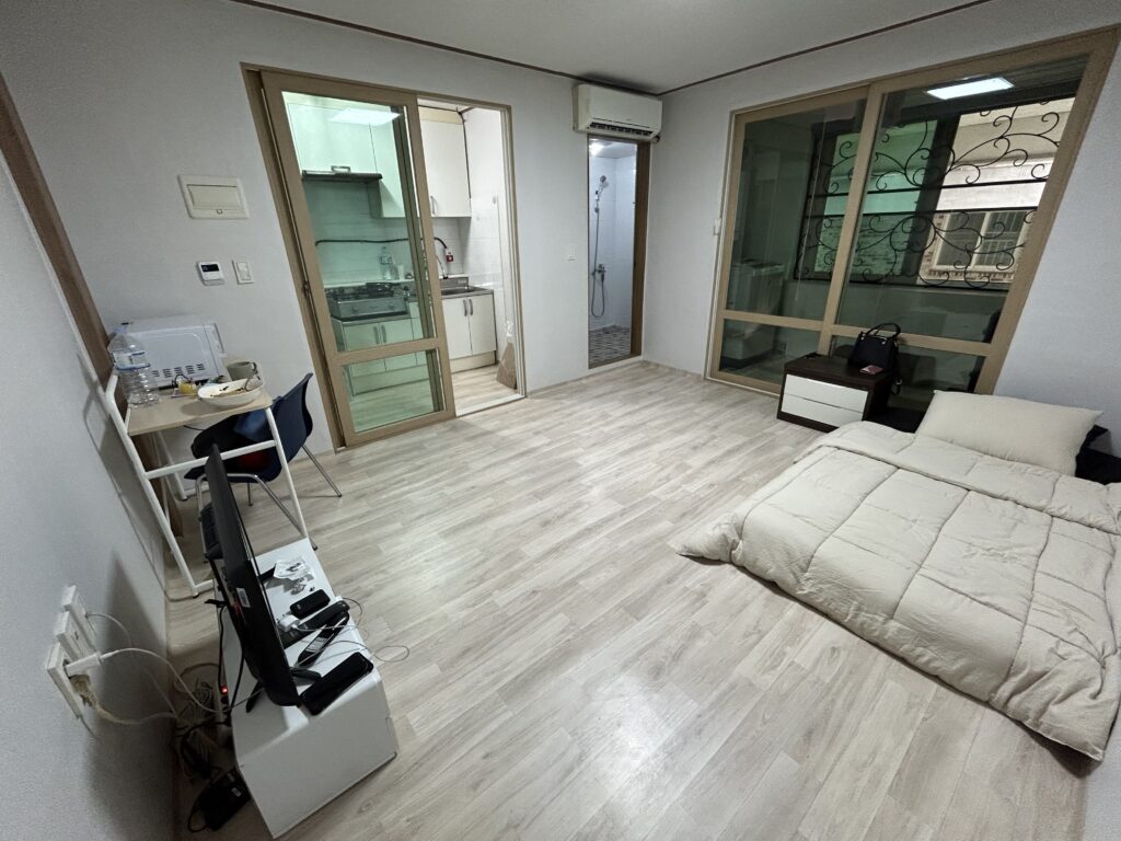 A living room in a Korean single apartment with a bed, table chair, TV and TV stand. One of the benefits to teach English in South Korea